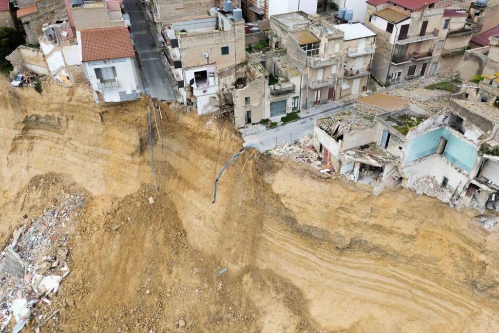 A deep hillside landslide next to densely built apartment blocks, exposing stratified soil and rubble at the edge of the cliff.