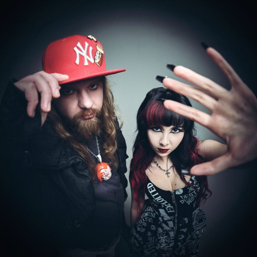 Two edgy individuals pose for a dramatic studio portrait: a bearded man in a red NY cap and necklace beside a woman with black and red hair and bold makeup, a hand reaching toward the camera in the foreground.