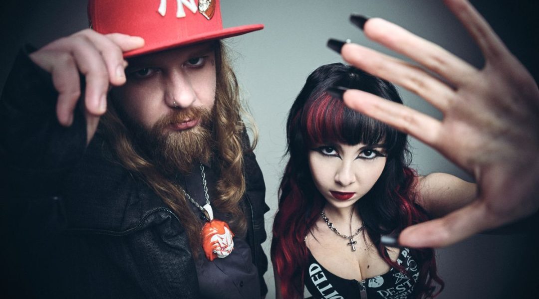 Two edgy individuals pose for a dramatic studio portrait: a bearded man in a red NY cap and necklace beside a woman with black and red hair and bold makeup, a hand reaching toward the camera in the foreground.